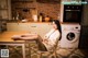 A woman sitting at a table in a kitchen next to a washing machine.