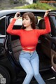 A woman in a red top and jeans leaning out the door of a car.