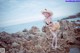 A woman in a bikini and hat standing on a rocky beach.