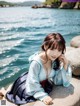 A woman in a blue dress sitting on a rock by the water.