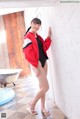 A woman in a red jacket and black bathing suit leaning against a wall.